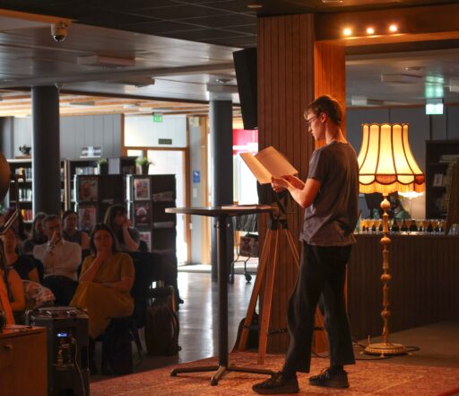 Male performer with glasses wearing a headset reads sheet music on a stand while an audience sits in a library-like space.