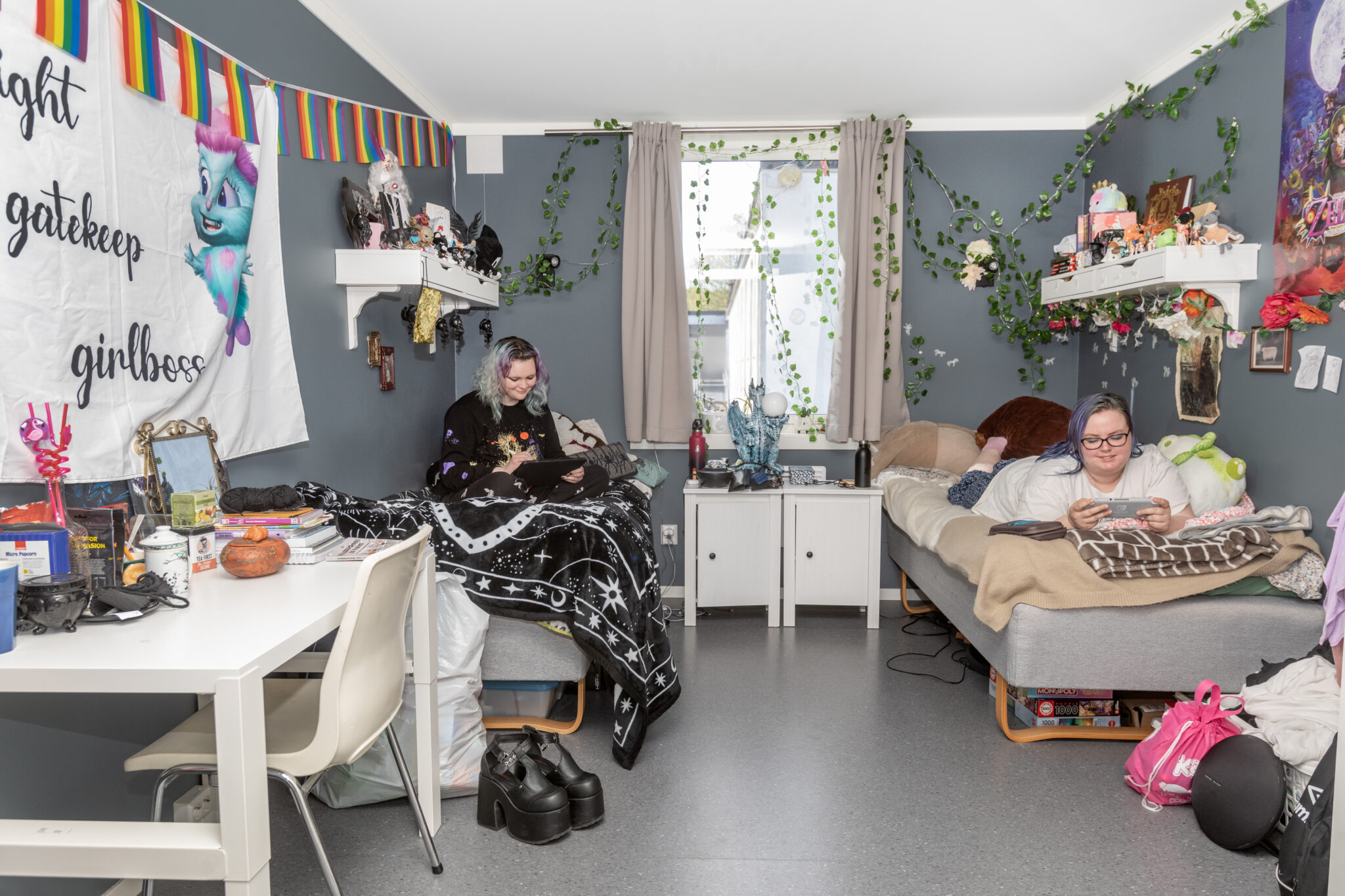 Two young women sit on separate beds in a cluttered, plant-filled dorm room, each using a device (tablet and phone). Decorations include rainbow bunting, ivy, and a large wall banner.