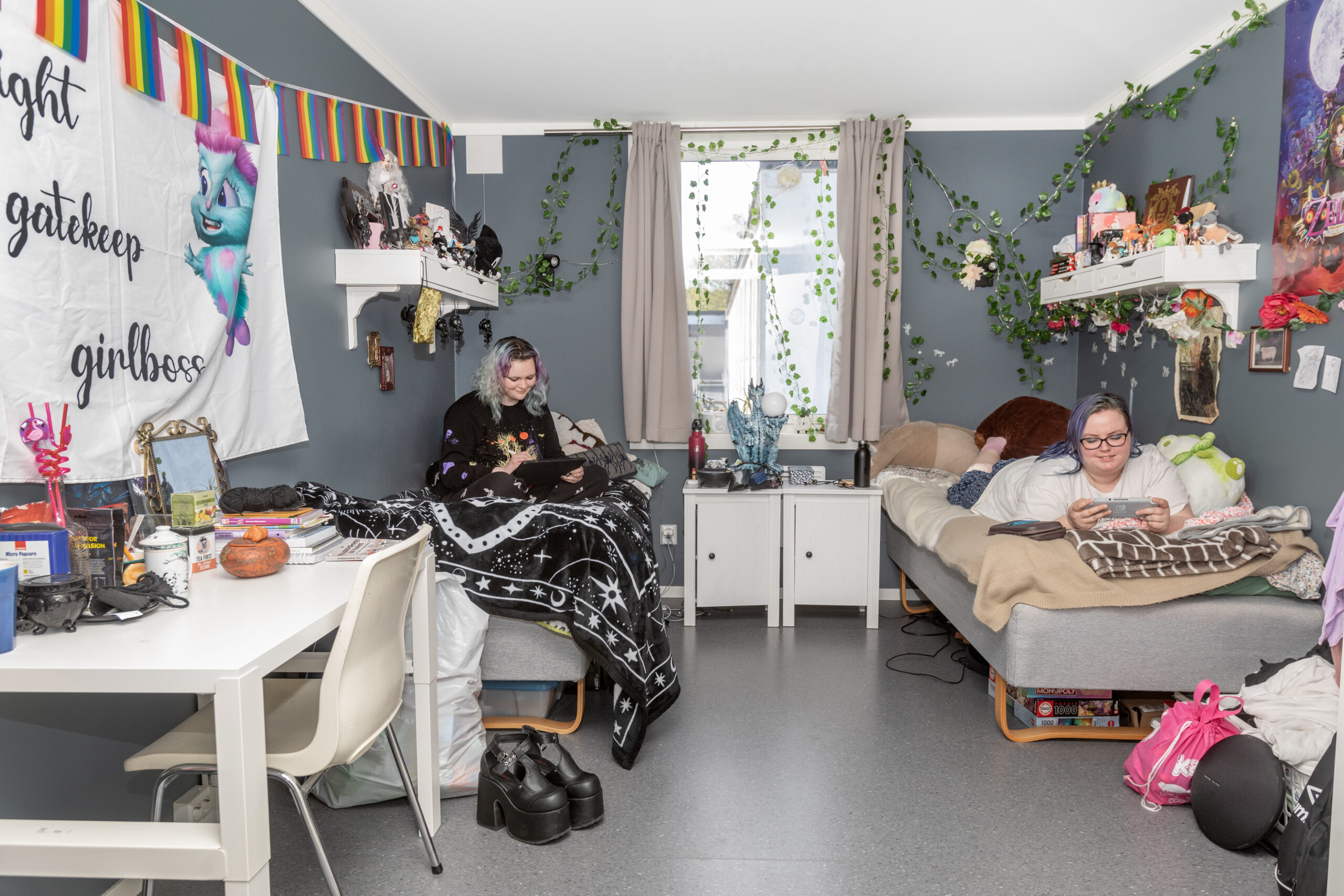Two young women sit on separate beds in a cluttered, plant-filled dorm room, each using a device (tablet and phone). Decorations include rainbow bunting, ivy, and a large wall banner.