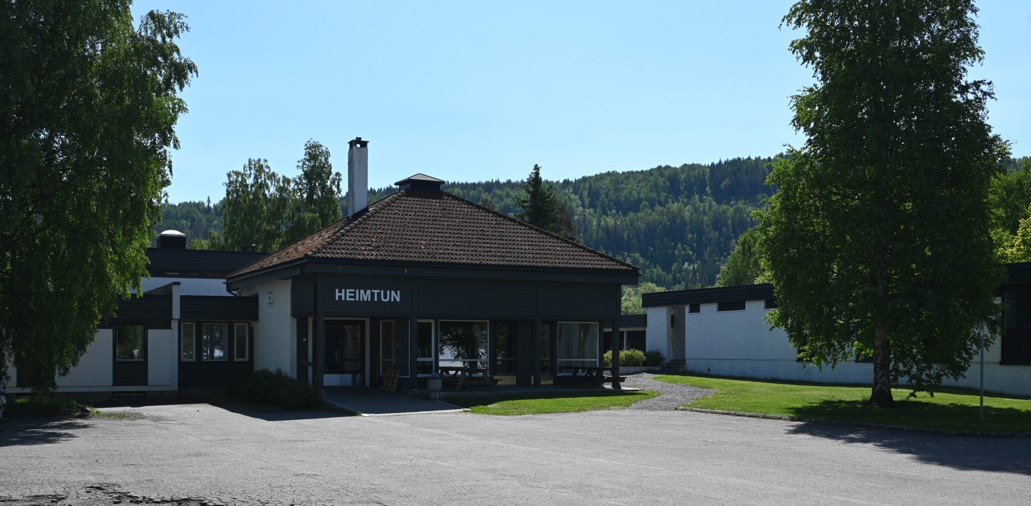 Front view of a small, dark facade building with a red tile roof and the sign 'HEIMTUN', set among green trees and a forested hillside behind.